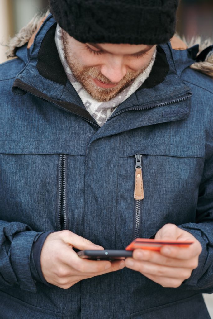 man in winter attire looking down at phone and credit card