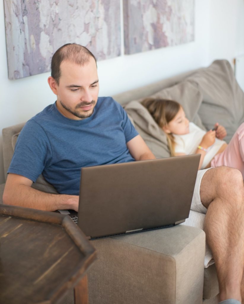 man using laptop while sitting next to daughter on sofa