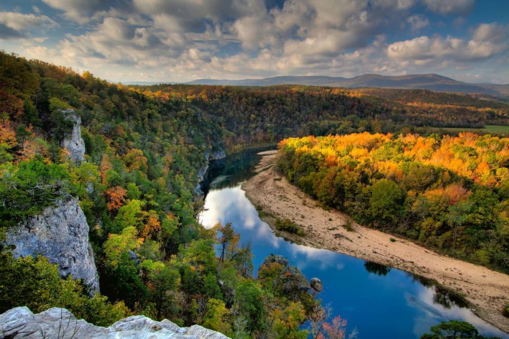 Buffalo National River meanders through a colorful autumn forest, with cliffs on one side and a backdrop of mountains and a cloudy sky.