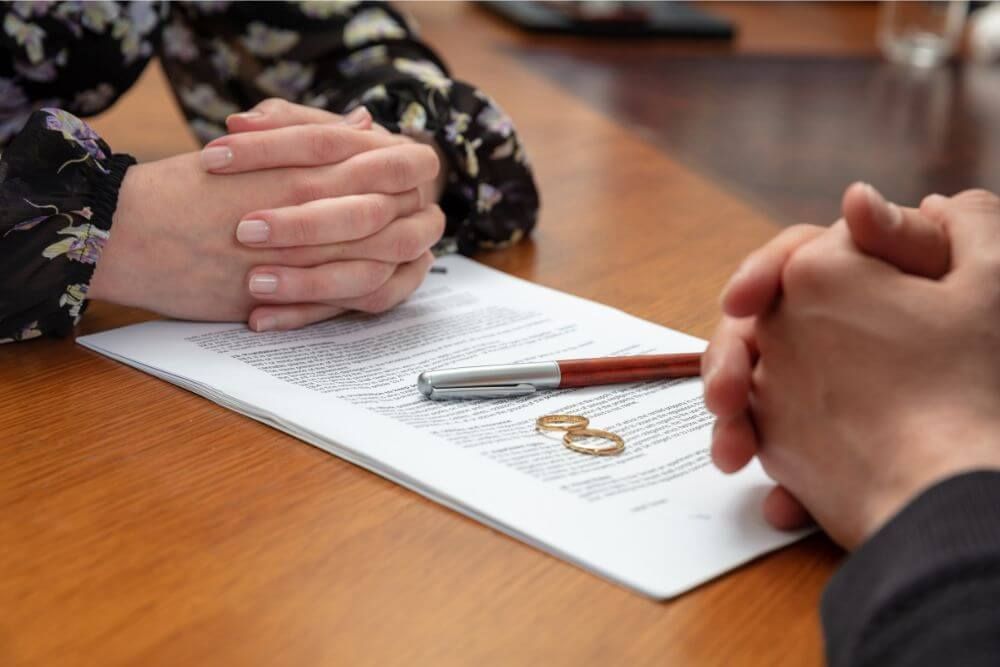 Two hands clasped together on top of a document, with wedding rings and a pen placed beside it, indicating a setting of a formal agreement or contract, likely a marriage.