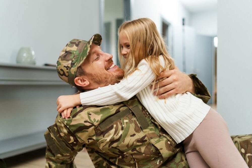 A smiling man in camouflage military attire embraces a joyful young girl in an indoor setting. Their affectionate interaction conveys a warm emotional connection.