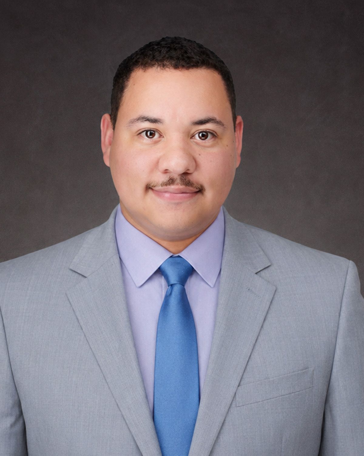 Professional headshot of a man in a light gray suit and blue tie against a dark backdrop.