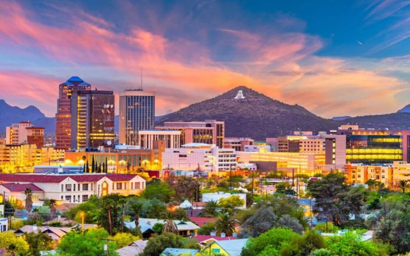 A cityscape at sunset with vibrant skies, featuring high-rise buildings, lit windows, and a mountain with a visible letter 'A' in the backdrop.