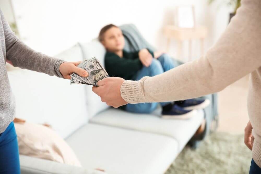 Two individuals exchange money, with one handing cash to the other, against a blurred indoor backdrop where a third person relaxes on a sofa.