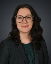 Portrait of a woman with wavy dark hair, tortoiseshell glasses, wearing a dark blazer and green blouse, smiling at the camera in a neutral studio background.