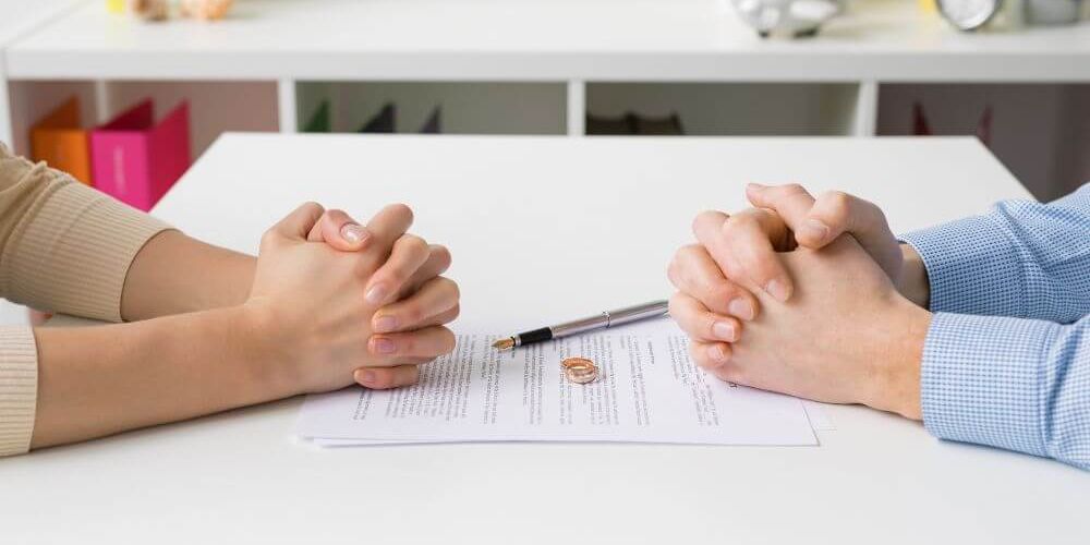 Two people are holding hands across a table with a marriage dissolution document between them, suggesting a couple going through a divorce.