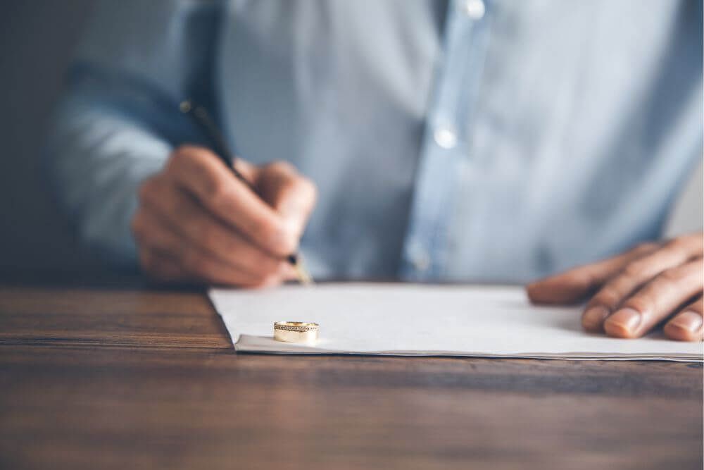 A person in a blue shirt is writing on paper, a gold wedding ring prominently in the foreground on a wooden table, implying a personal or significant moment.