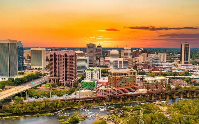 A skyline displaying numerous buildings at sunset, with a river flowing in the foreground and verdant trees peppered throughout the urban landscape.