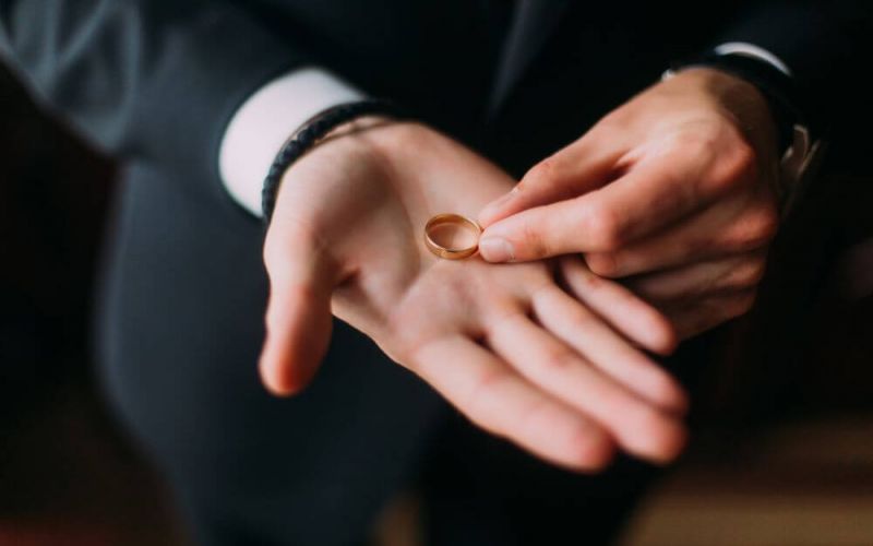 A person in a dark suit holds a golden wedding ring delicately between two fingers, likely symbolizing a moment related to a marriage ceremony.