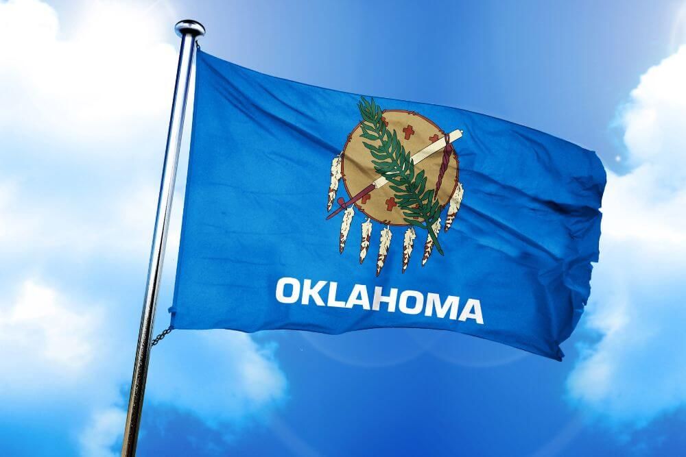 A blue Oklahoma state flag flutters on a flagpole against a backdrop of a clear sky with a few clouds.