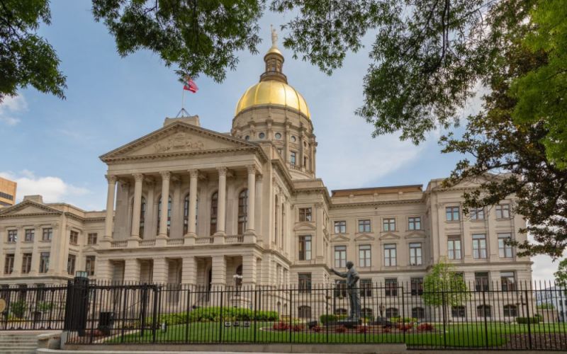 Georgia capital building with a golden dome stands prominently under a blue sky, framed by lush foliage in a city environment.