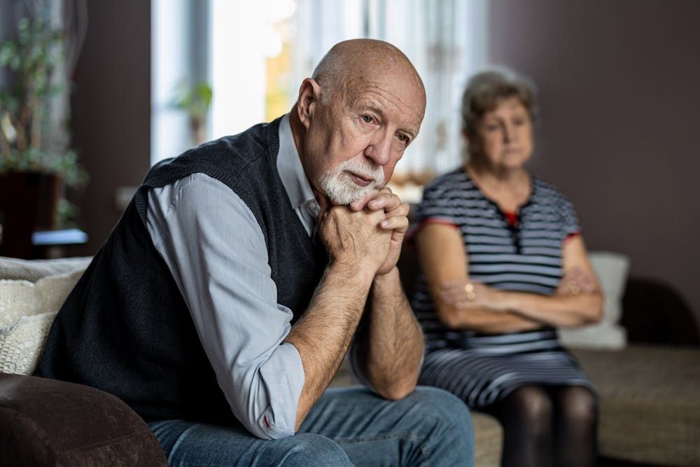 An elderly man sits pensively with his hand under his chin, his gaze directed off-camera, while a woman sits behind him, arms crossed, looking slightly away, suggesting tension. They are in a cozy home setting.