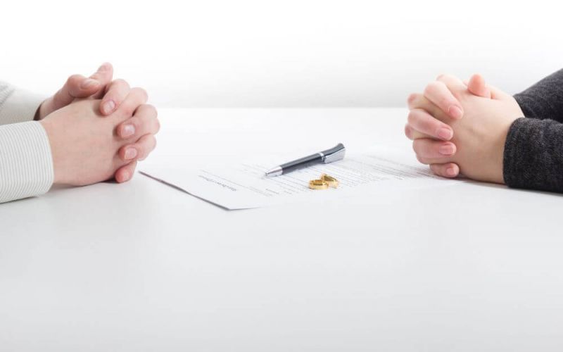 Two people's hands clasped together on a table, with a document, pen, and two wedding rings between them, suggesting a context of either marriage or divorce proceedings.