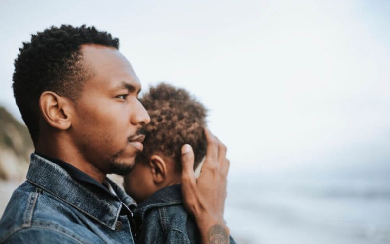 A man gently holds a child, both in denim attire, against a blurred coastal backdrop. The mood appears contemplative and serene.