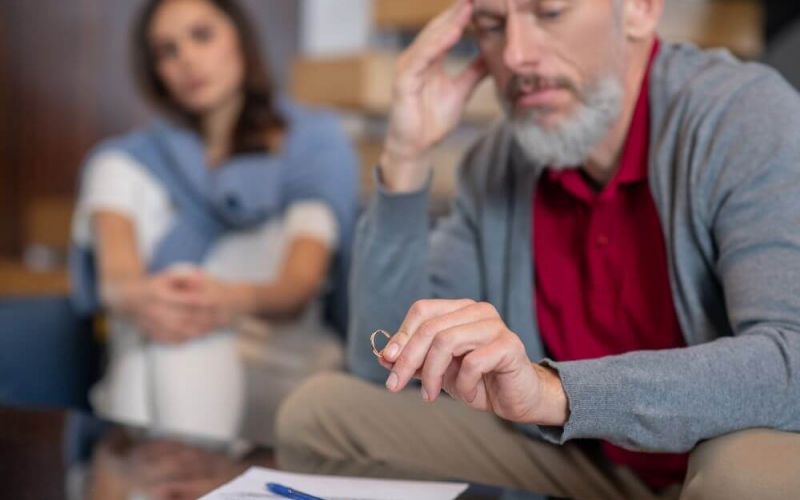 A man looks contemplatively at a wedding ring, appearing distressed, with a blurred woman in the background, suggesting a context of relationship strain or decision-making.