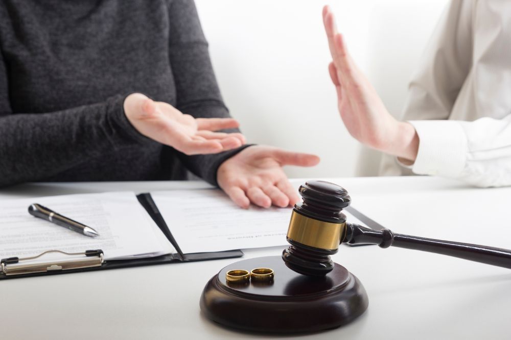 Two people gesticulate during a discussion, with a gavel and wedding rings on the table, suggesting a divorce proceeding context.