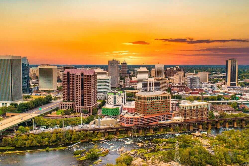 A skyline displaying numerous buildings at sunset, with a river flowing in the foreground and verdant trees peppered throughout the urban landscape.