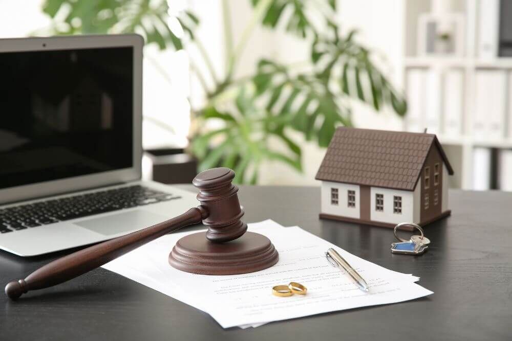 A wooden gavel rests on a document beside a laptop, house model, and keys, implying legal proceedings, possibly real estate, on an office desk.