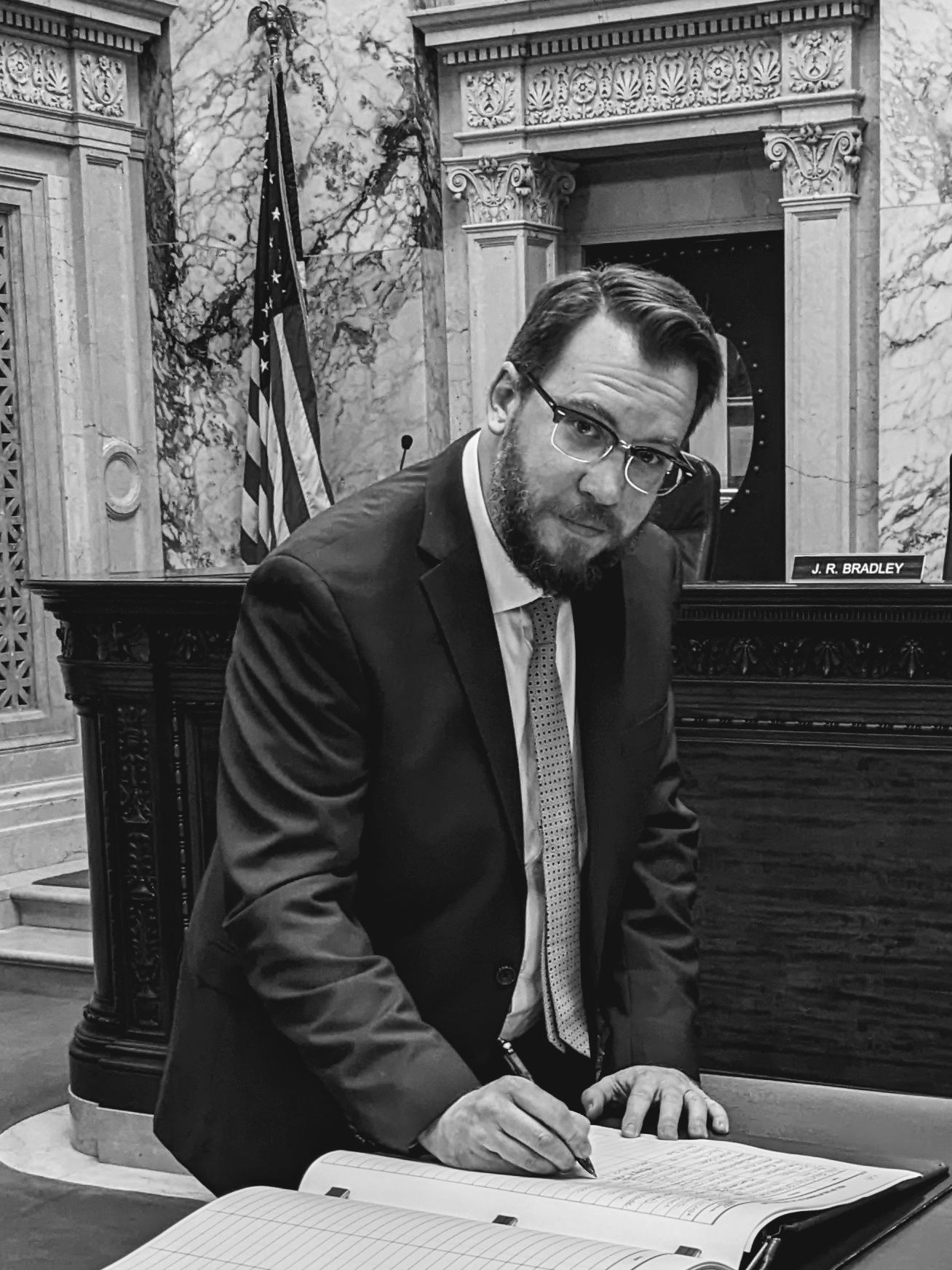 Un hombre con traje y gafas firma un libro de contabilidad en un escritorio en una cámara de mármol, con una bandera estadounidense al fondo y una placa con el nombre JR BRADLEY cerca.