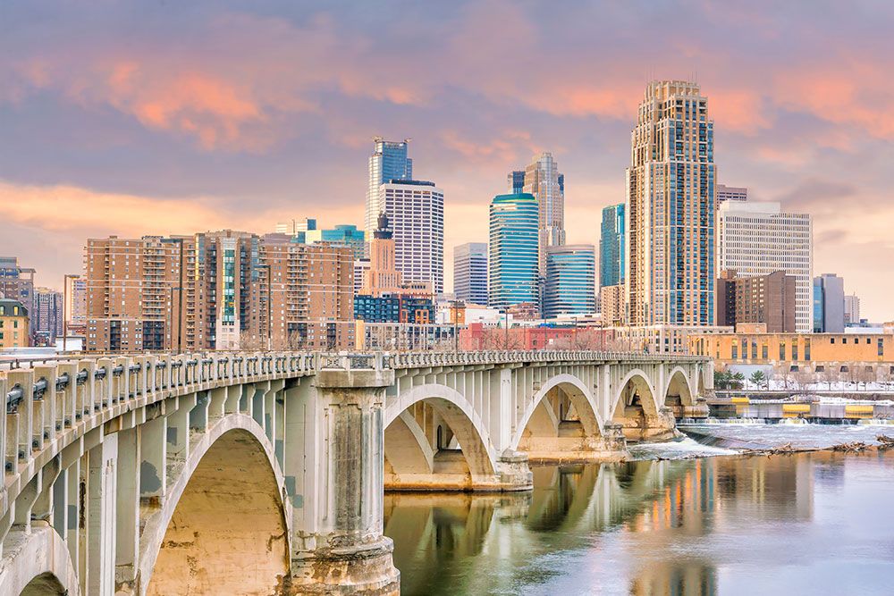 Stone arch bridge in Minneapolis Minnesota spans a calm river, leading towards a metropolitan skyline with skyscrapers under a pastel sunset sky.