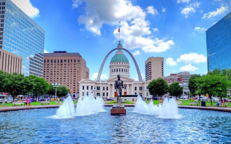 Water fountains gush in a park with people, framed by a domed building and the arching Gateway Arch in a vibrant urban landscape.