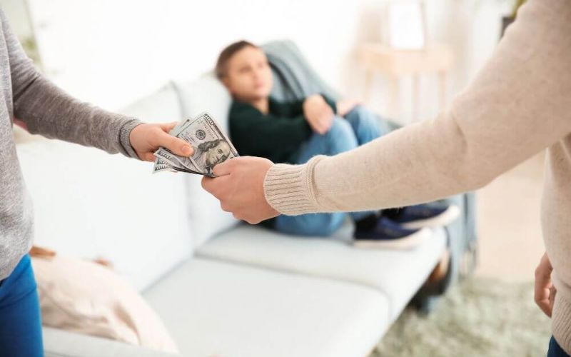 Two individuals exchange money, with one handing cash to the other, against a blurred indoor backdrop where a third person relaxes on a sofa.
