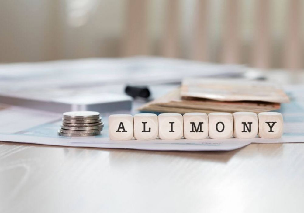 Stacked coins and scrabble tiles spelling "ALIMONY" rest on a paper-filled desk, suggesting financial matters related to spousal support payments.