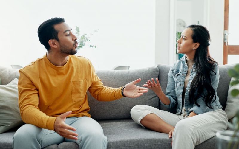 Two people are conversing on a grey couch, gesturing expressively, in a bright living room setting.