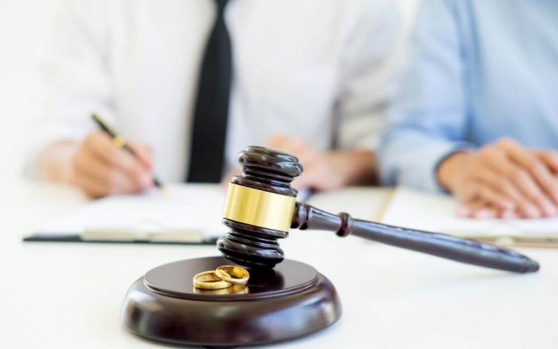 A gavel and two gold wedding rings on a sound block, with two people writing in the background, possibly depicting a legal setting focused on marital issues.