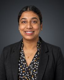 Professional headshot: smiling woman with short dark hair, gold earrings, wearing a black blazer and patterned blouse against a dark studio backdrop