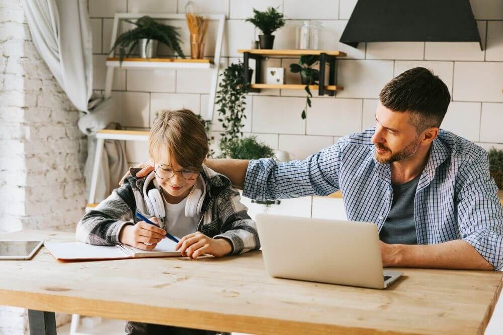 A young child writes in a notebook while an adult male observes and uses a laptop, both seated at a wooden table in a homey, bright room.