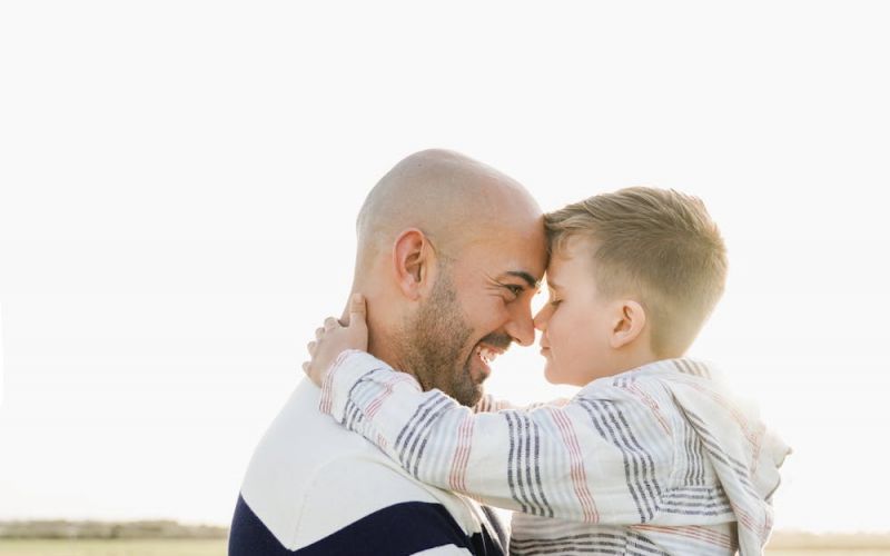 A man and a young boy are smiling and touching foreheads affectionately in a bright, open field. They appear joyful and are embracing closely.