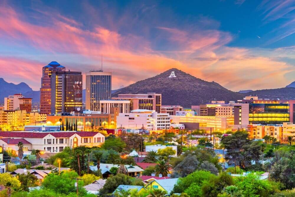 A cityscape at sunset with vibrant skies, featuring high-rise buildings, lit windows, and a mountain with a visible letter 'A' in the backdrop.