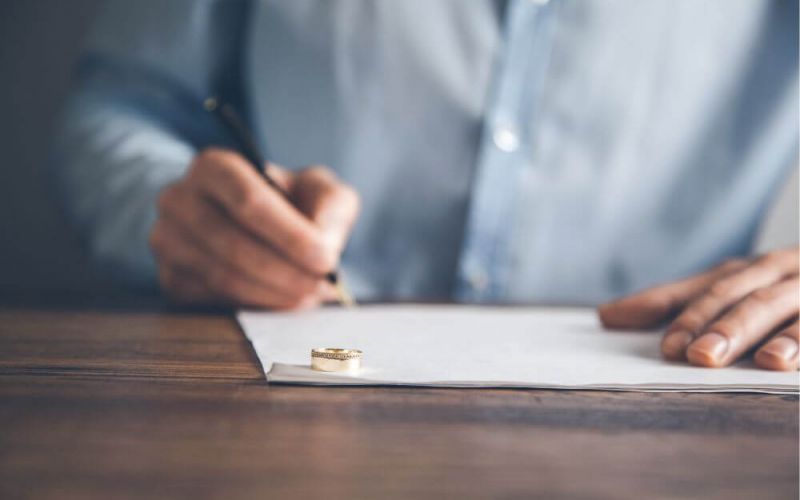 A person in a blue shirt is writing on paper, a gold wedding ring prominently in the foreground on a wooden table, implying a personal or significant moment.