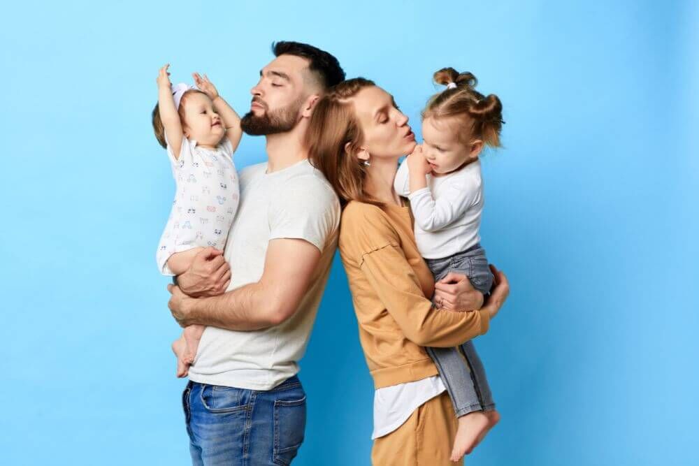 A man and a woman each hold a child, while standing and looking affectionate against a plain blue background.