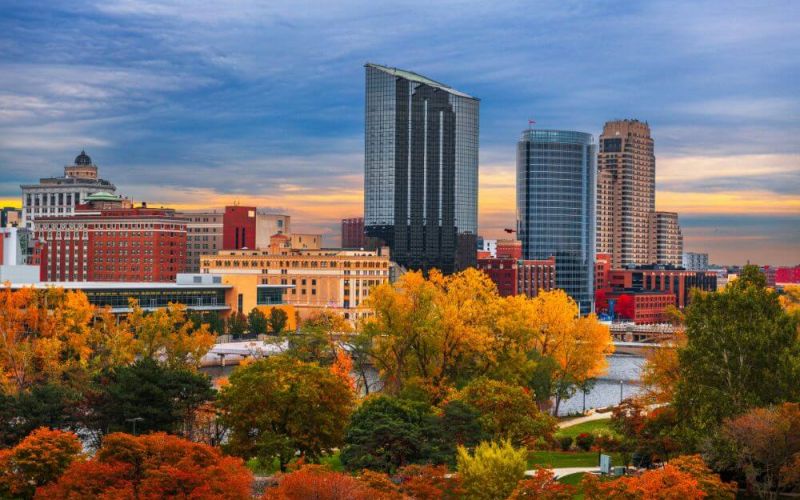 Skyscrapers tower over an urban park with autumn-colored trees, beside a calm river, under a dusky sky with soft clouds.