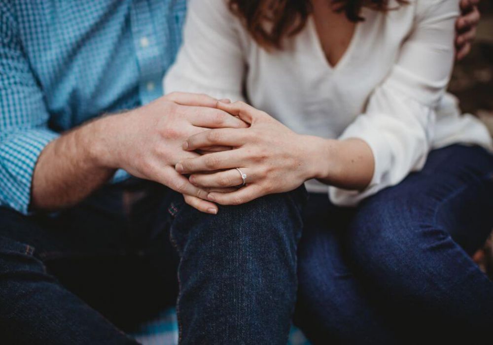 Two people, likely a couple, are sitting close together with their hands clasped on their laps, showcasing a diamond ring on the woman's finger, suggesting engagement.