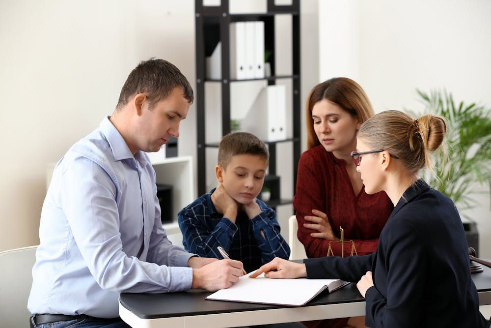 Four people are in an office setting, with one person signing a document. There’s a sense of consultation or agreement being formalized.