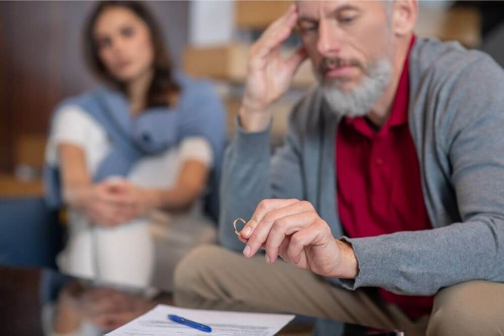 A man looks contemplatively at a wedding ring, appearing distressed, with a blurred woman in the background, suggesting a context of relationship strain or decision-making.