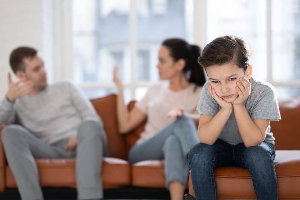 A distressed boy sits foreground with hands on cheeks, while a man and woman argue in the background on a couch, in a bright living room.