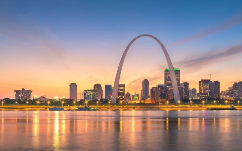 The St. Louis Arch looms over a skyline at dusk, with the city's lights reflecting on the tranquil waters of a river.