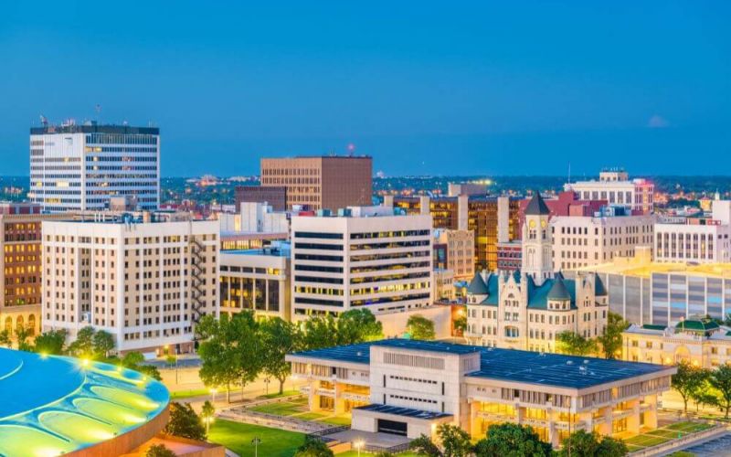 A cityscape at dusk with illuminated buildings against a twilight sky, featuring a mix of modern and historic architecture, and a clear foreground park area.