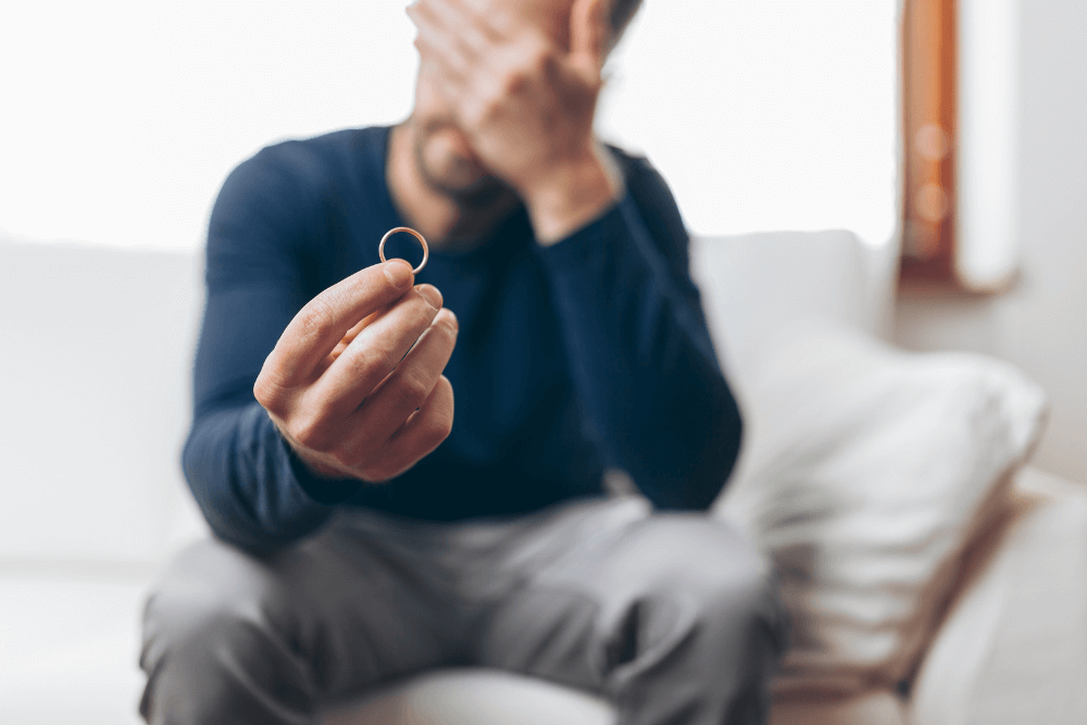 Man sitting on a couch holding up a wedding ring that is in focus