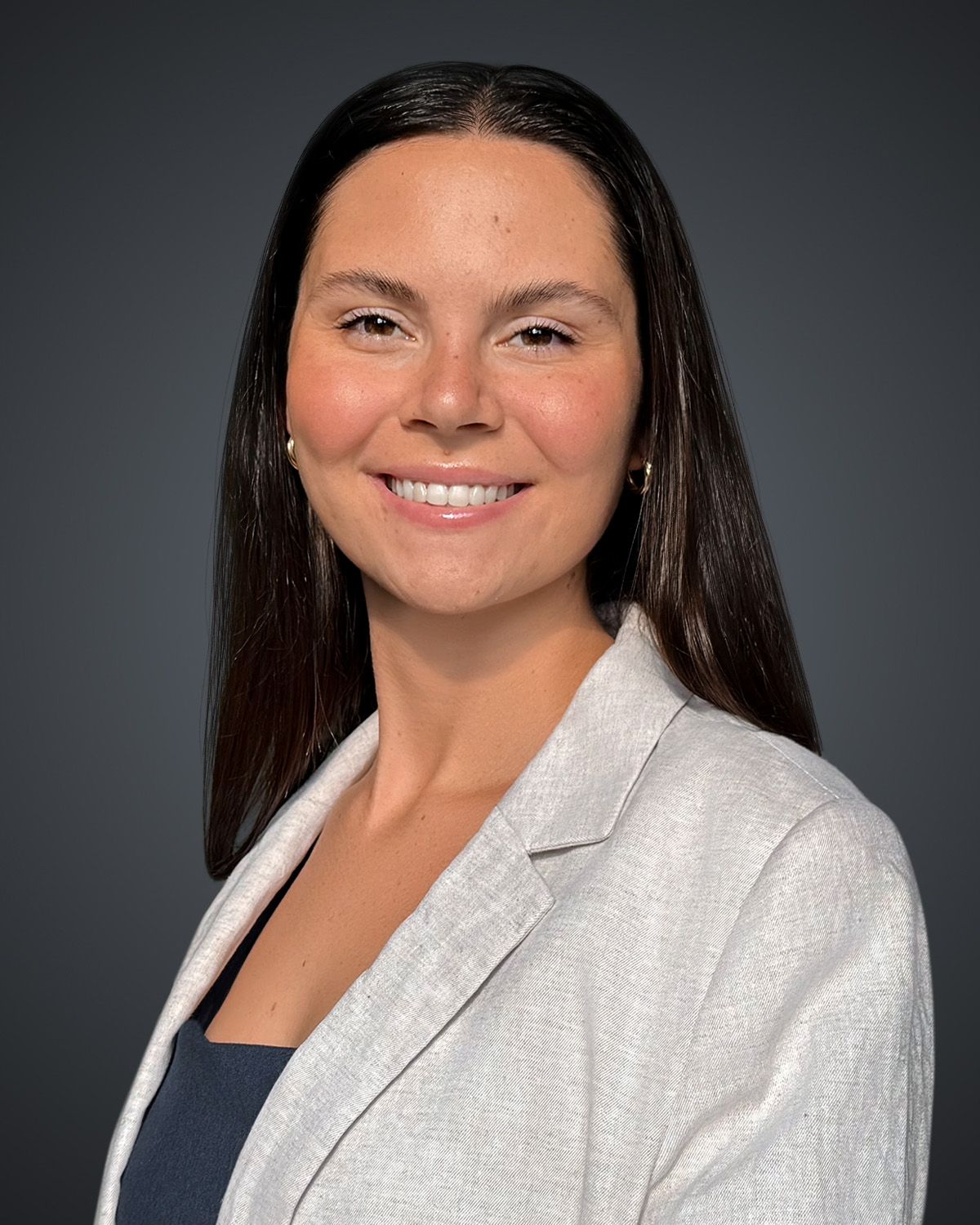 Professional headshot: smiling woman with long dark hair, wearing a light-gray blazer and navy top against a dark gradient background.