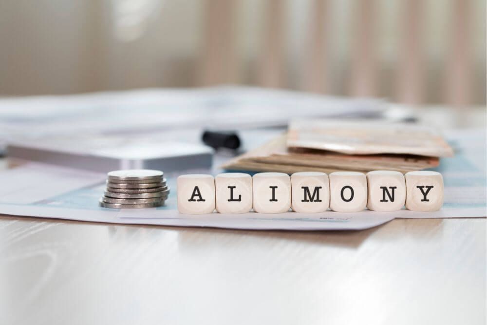 Stacked coins and scrabble tiles spelling "ALIMONY" rest on a paper-filled desk, suggesting financial matters related to spousal support payments.