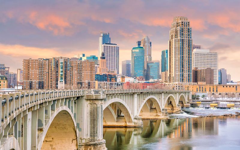 Stone arch bridge in Minneapolis Minnesota spans a calm river, leading towards a metropolitan skyline with skyscrapers under a pastel sunset sky.