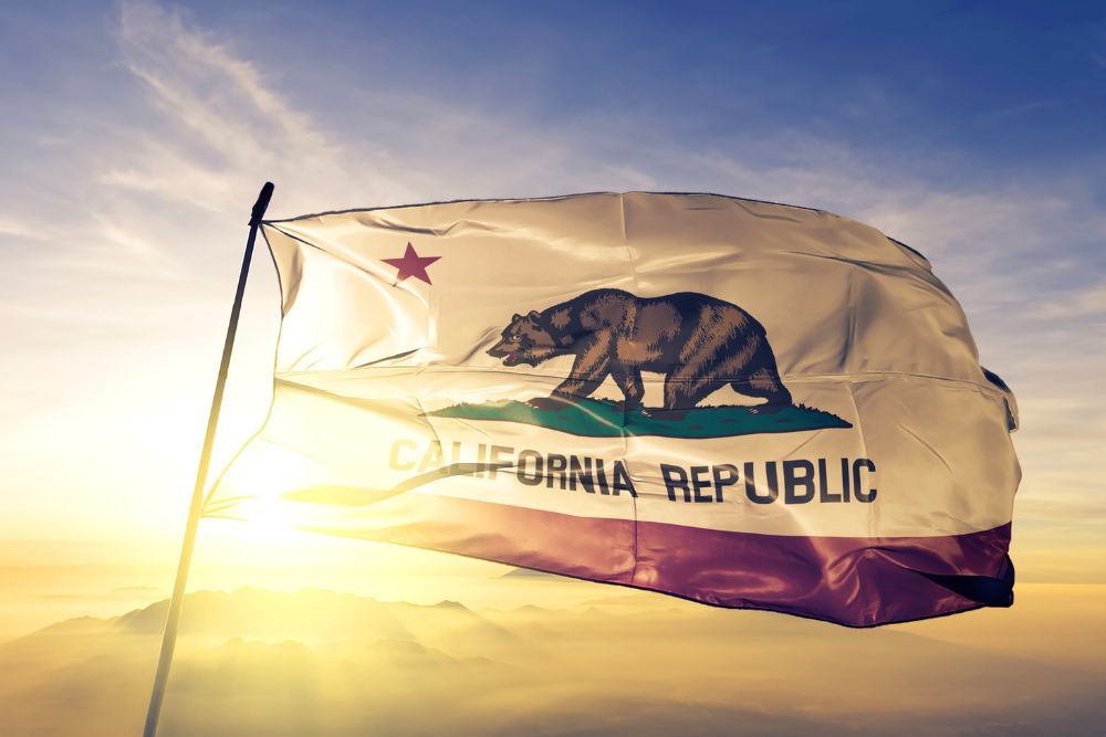 A California state flag flutters in the breeze against a backdrop of hazy sunrise and clouds.