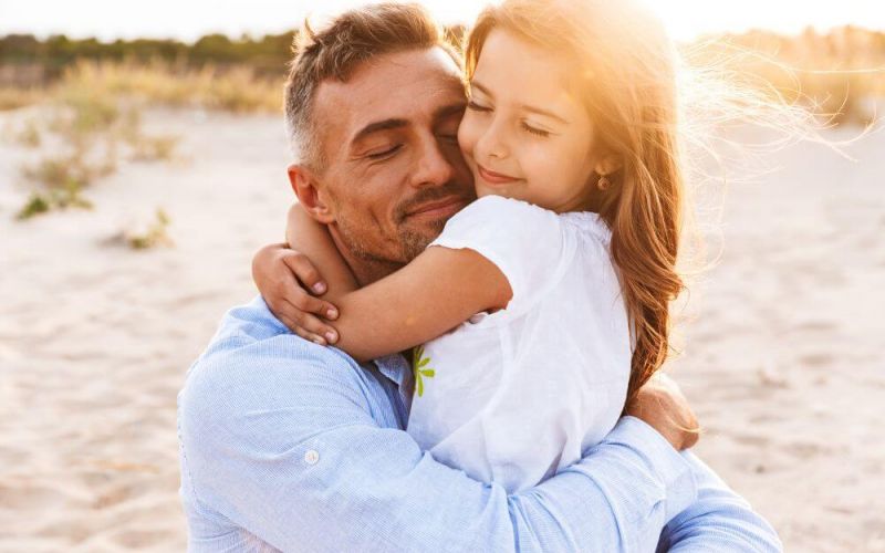 A man embraces a young girl affectionately on a sandy beach at sunset. Both exhibit a content and peaceful demeanor.