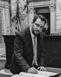 Man in a suit and glasses signs a ledger at a desk in a marble chamber, with a U.S. flag in the background and a nameplate nearby reading J. R. BRADLEY.