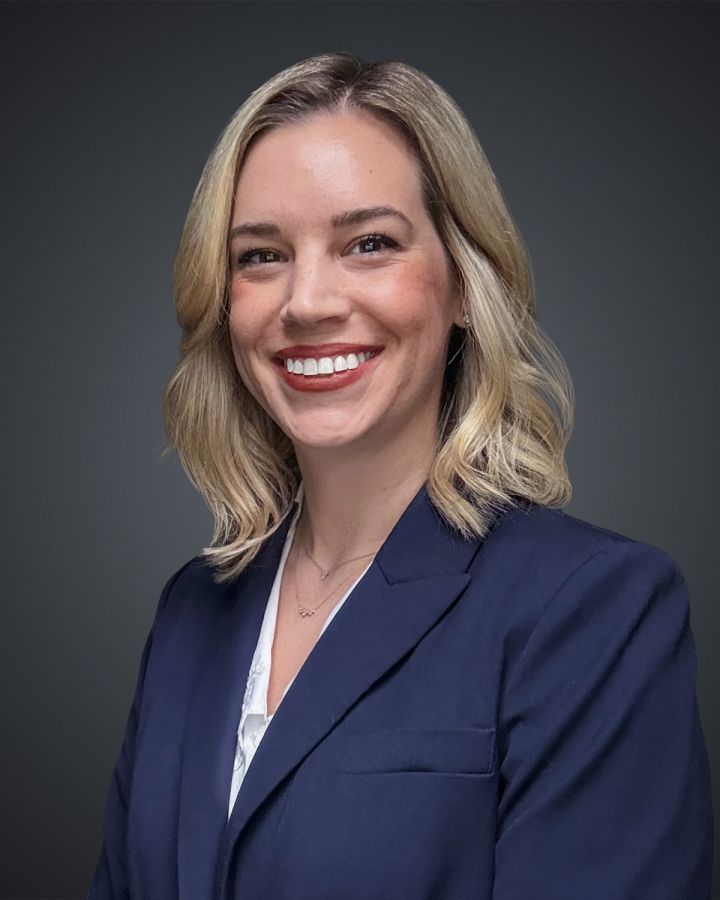 Portrait of a smiling woman with blonde hair in a navy blazer against a dark gradient background.